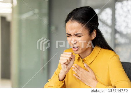 Young woman suffering from a cough and sore throat, showing symptoms of flu or cold in an office environment, experiencing discomfort and pain from sickness Young woman suffering from a cough and sore throat, showing symptoms of flu or cold in an office environment, experiencing discomfort and pain from sickness 137544779
