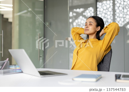 Female professional taking a mindful moment, relaxing with her eyes closed and hands behind head, enjoying stress relief during a break at her modern office workspace Female professional taking a mindful moment, relaxing with her eyes closed and hands behind head, enjoying stress relief during a break at her modern office workspace 137544783