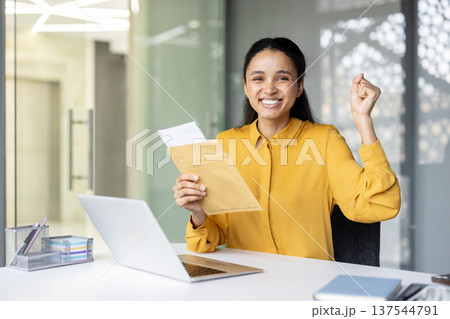 Happy young multiracial business woman celebrating achievement, expressing excitement while holding an important letter in an envelope at her modern office desk with a laptop Happy young multiracial business woman celebrating achievement, expressing excitement while holding an important letter in an envelope at her modern office desk with a laptop 137544791