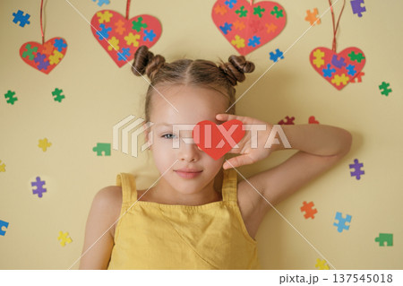 A beautiful, kind girl with clear eyes on yellow background with puzzle pieces hides eyes behind hand, demonstrating red heart. A symbol of support, care and universal love for people with autism 137545018