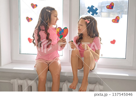 A girl gives a heart-shaped postcard with puzzles to her sister with autism spectrum disorder. The children's room and window glass are decorated with pieces of multicolored puzzles for Autism Day A girl gives a heart-shaped postcard with puzzles to her sister with autism spectrum disorder. The children's room and window glass are decorated with pieces of multicolored puzzles for Autism Day 137545046