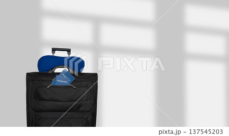 Black rolling suitcase with blue neck pillow and travel tag positioned against a light gray wall with soft shadows creating a minimalist travel-themed setting 137545203