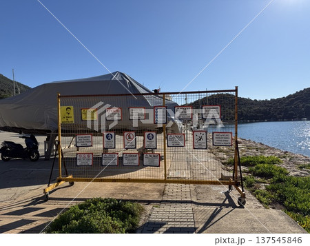 Warning signs on fenced area near water and covered boat, clear sky and nature in background Warning signs on fenced area near water and covered boat, clear sky and nature in background 137545846