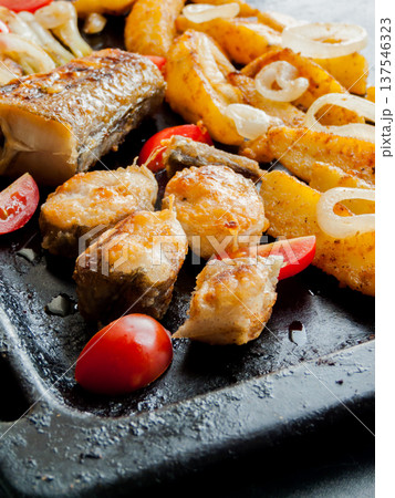 Oven pan with sliced baked potatoes, fish steaks and vegetables with onion on table. Oven pan with sliced baked potatoes, fish steaks and vegetables with onion on table. 137546323