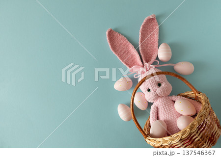Woven basket with pink crocheted bunny doll surrounded by pastel Easter eggs against soft blue background Woven basket with pink crocheted bunny doll surrounded by pastel Easter eggs against soft blue background 137546367