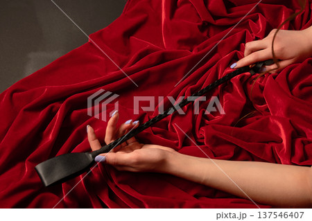 Female hands with manicured nails holding a black leather riding crop on luxurious red velvet fabric. 137546407