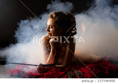 Elegant woman in shibari rope tie holding a riding crop, lying on red velvet in dramatic smoke. 137546408