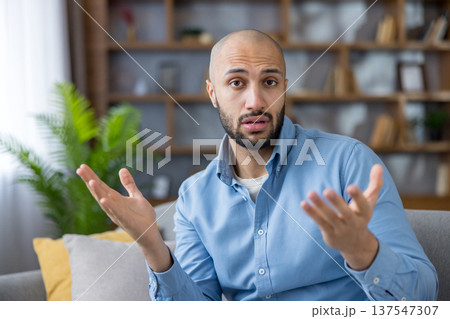 Young man sitting on a sofa, looking at the camera with a puzzled expression and gesturing with open hands, conveying doubts and seeking an explanation in a home setting 137547307
