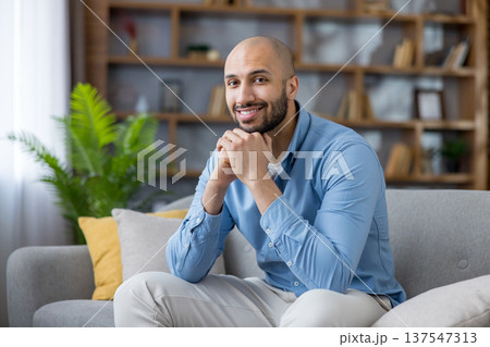 Smiling bald man with beard sitting comfortably on a sofa in his living room, looking directly at the camera with a confident, relaxed expression, enjoying leisure time Smiling bald man with beard sitting comfortably on a sofa in his living room, looking directly at the camera with a confident, relaxed expression, enjoying leisure time 137547313