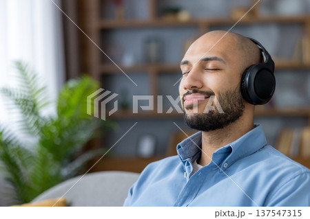 Man listening to audio with eyes closed, finding peace and enjoying a moment of relaxation while embracing sound therapy in a comfortable home interior Man listening to audio with eyes closed, finding peace and enjoying a moment of relaxation while embracing sound therapy in a comfortable home interior 137547315