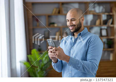 Smiling bearded bald man in a blue shirt using his smartphone indoors, happily texting and browsing, illustrating modern digital communication, connectivity and casual work-life lifestyle 137547321