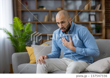 Man experiencing acute chest pain while sitting on a sofa in his living room, struggling with a potential heart attack or respiratory issues, needing immediate medical attention Man experiencing acute chest pain while sitting on a sofa in his living room, struggling with a potential heart attack or respiratory issues, needing immediate medical attention 137547336
