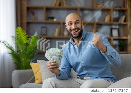 Young man on couch holding cash and pumping his fist, celebrating financial success and joy at home with a big smile and excited, triumphant expression 137547347