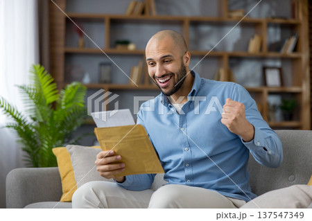Excited man at home celebrates good news after opening a mail letter, sitting on a sofa with fist raised, smiling in relief and joy over an approval or success notification Excited man at home celebrates good news after opening a mail letter, sitting on a sofa with fist raised, smiling in relief and joy over an approval or success notification 137547349