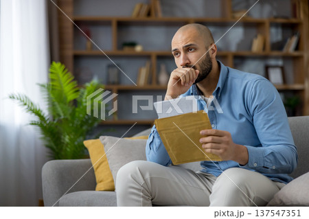 Man feeling anxious and stressed while holding an open envelope with a letter, contemplating financial problems, debt, or an unemployment notice, sitting on a sofa in his modern living room 137547351