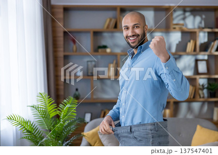 Man proudly celebrating his successful weight loss journey, clenching a fist in triumph and showing off the loose fit of his old, oversized jeans at home 137547401