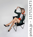 An elegant white woman in a green business suit talks on a red telephone while sitting on a chair against the backdrop of an isolated space. Office. Curly hair, brown-haired 137552473