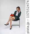 An elegant white woman in a green business suit talks on a red telephone while sitting on a chair against the backdrop of an isolated space. Office. Curly hair, brown-haired 137552474