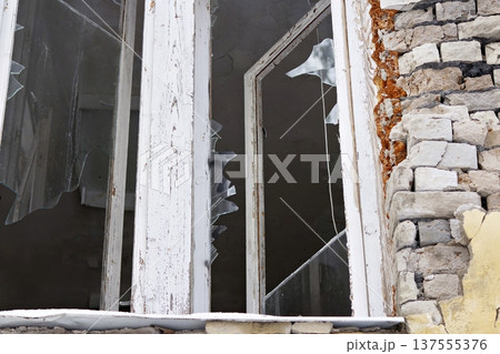 Dilapidated window frame with shattered glass panes, peeling white paint, and crumbling brickwork revealing rusty insulation 137555376