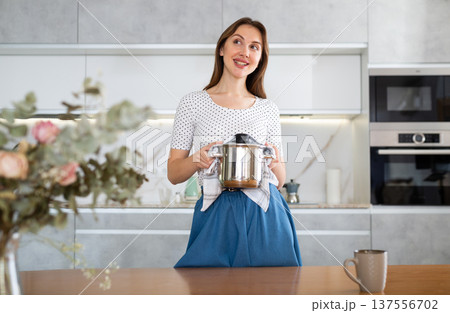 Smiling girl is standing in kitchen in casual clothes and holding stainless steel pot in hands. Smiling girl is standing in kitchen in casual clothes and holding stainless steel pot in hands. 137556702