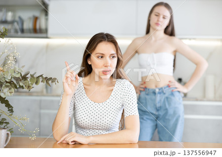 Angry mother sitting at the kitchen table, daughter standing behind Angry mother sitting at the kitchen table, daughter standing behind 137556947