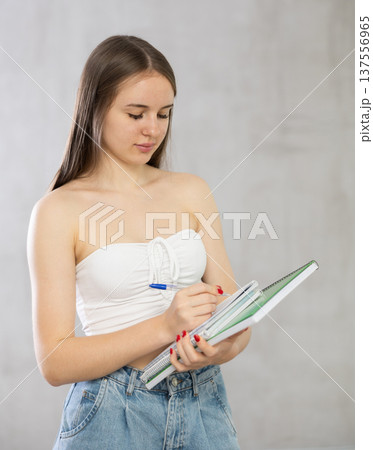Girl stands and holding stack of notebooks in one hand, and writes information in diary. 137556965