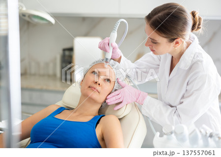 Happy adult woman getting facial deep hydration procedure from experienced cosmetologist in medicine office. Concept of professional cosmetology and rejuvenation Happy adult woman getting facial deep hydration procedure from experienced cosmetologist in medicine office. Concept of professional cosmetology and rejuvenation 137557061