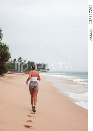 Beach Run Alone. Morning Jog Along Sandy Shore. Lone Woman Exercises On Beach With Footprints In Sand 137557260