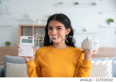 Young Asian woman holding piggy bank and calculator sitting on couch in living room at home Young Asian woman holding piggy bank and calculator sitting on couch in living room at home 137558582
