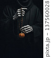 Skeleton hands in black hooded robe holding dark prayer beads with small carved Halloween pumpkin jack-o-lantern. 137560028