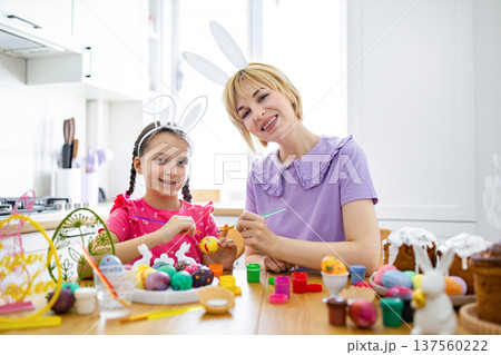 A mother and daughter happily paint colorful Easter eggs together at a kitchen table, wearing bunny ears and surrounded by festive decorations A mother and daughter happily paint colorful Easter eggs together at a kitchen table, wearing bunny ears and surrounded by festive decorations 137560222