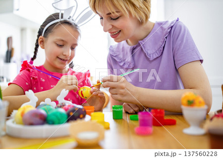 A mother and her daughter are happily painting colorful Easter eggs together at a wooden table, creating festive decorations for the holiday 137560228