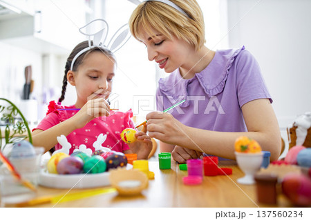 A mother and her daughter wearing bunny ears are happily painting colorful Easter eggs together at a wooden table with paints and brushes 137560234