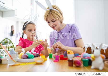 A mother and her daughter wearing bunny ears are happily painting colorful Easter eggs together at a wooden table, surrounded by art supplies and Easter cakes 137560236