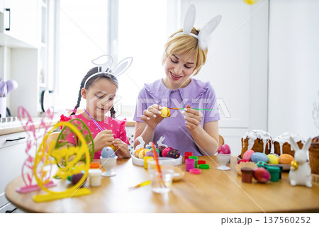 A mother and her daughter wearing bunny ears are happily painting colorful Easter eggs together at a wooden table, surrounded by festive decorations and cakes A mother and her daughter wearing bunny ears are happily painting colorful Easter eggs together at a wooden table, surrounded by festive decorations and cakes 137560252
