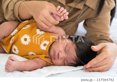 Newborn baby in mustard yellow onesie sleeping on white bed while mother hand holding baby girl tiny fingers with love Newborn baby in mustard yellow onesie sleeping on white bed while mother hand holding baby girl tiny fingers with love 137564792