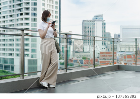 solo asian woman wear protective mask during use phone and relax at rooftop with city background 137565892