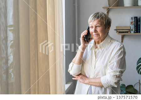 An elderly happy woman enjoys a phone conversation by the window, chatting with loved ones and reflecting on home life An elderly happy woman enjoys a phone conversation by the window, chatting with loved ones and reflecting on home life 137571570