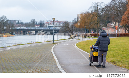 A man walks along a riverside path with a bike trailer stroller in Germany. The scene is set in a park with trees and a bridge in the background. A man walks along a riverside path with a bike trailer stroller in Germany. The scene is set in a park with trees and a bridge in the background. 137572083