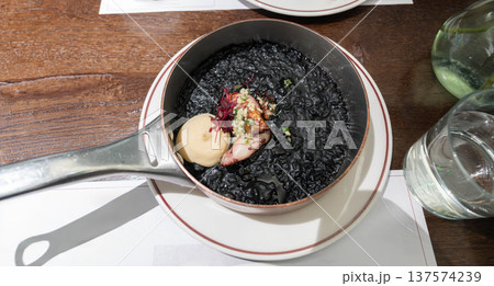 Mediterranean squid ink rice served in a metal pan with tender squid and creamy sauce, styled as a rich black seafood dish with elegant restaurant presentation. Mediterranean squid ink rice served in a metal pan with tender squid and creamy sauce, styled as a rich black seafood dish with elegant restaurant presentation. 137574239