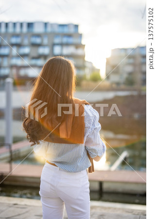 Elegant young woman in old money style standing by riverside in Prague during golden hour, back view portrait. Quiet luxury lifestyle and refined European femininity concept. Elegant young woman in old money style standing by riverside in Prague during golden hour, back view portrait. Quiet luxury lifestyle and refined European femininity concept. 137575102