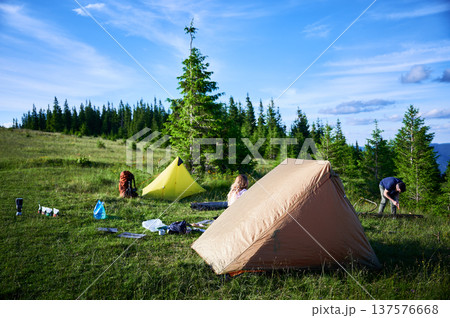 Picturesque mountain campsite on grassy hill with tents. Camper and child tend to campfire, surrounded by backpacks and gear. Rolling hills, evergreens, clear blue sky. Peaceful outdoor adventure. 137576668