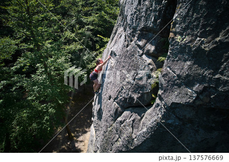 Aerial view of woman rock climber ascending rugged limestone cliff with harness and rope for safety. Sportswoman climbing on vertical large boulder at Dobvush Rocks in Carpathian mountains, Ukraine. Aerial view of woman rock climber ascending rugged limestone cliff with harness and rope for safety. Sportswoman climbing on vertical large boulder at Dobvush Rocks in Carpathian mountains, Ukraine. 137576669
