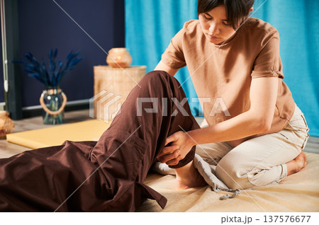 Masseuse making thai yoga massage. Woman therapist doing traditional massage treatment for female patient indoors. Client lying on mat, meditating, enjoying hand massage in calm atmosphere. 137576677