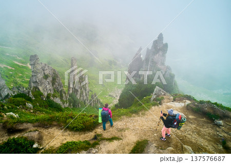 Aerial view of two hikers stand at edge of misty mountain ridge, surrounded by towering rock formations and lush green vegetation. One hiker taking photo while other looks out over foggy landscape. Aerial view of two hikers stand at edge of misty mountain ridge, surrounded by towering rock formations and lush green vegetation. One hiker taking photo while other looks out over foggy landscape. 137576687