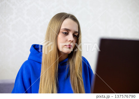 Young woman with straight blonde hair in a blue hoodie is seated indoors, concentrating on her laptop screen, with a light-patterned wall visible in the background Young woman with straight blonde hair in a blue hoodie is seated indoors, concentrating on her laptop screen, with a light-patterned wall visible in the background 137576790