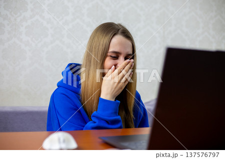 Blonde girl laughs at funny football moment on laptop, covering mouth with hand. Amusing sports blooper or unexpected play during match makes her giggle. Entertainment at home 137576797