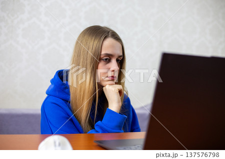 Blonde girl in blue hoodie rests chin on clenched fist while staring at laptop screen. Poor posture during online study session reflects learning difficulties and ergonomic challenges at home Blonde girl in blue hoodie rests chin on clenched fist while staring at laptop screen. Poor posture during online study session reflects learning difficulties and ergonomic challenges at home 137576798