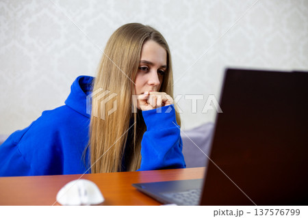 Young blonde woman in blue hoodie nervously bites her fist while watching live sports on laptop. Tense moment during crucial game makes her anxious for her favorite team. Intense emotions at home Young blonde woman in blue hoodie nervously bites her fist while watching live sports on laptop. Tense moment during crucial game makes her anxious for her favorite team. Intense emotions at home 137576799