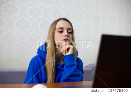 Young woman of Caucasian descent in a blue hoodie rests her chin on her hand while looking at a laptop screen, with a light-patterned wall and wooden table visible in the background Young woman of Caucasian descent in a blue hoodie rests her chin on her hand while looking at a laptop screen, with a light-patterned wall and wooden table visible in the background 137576800